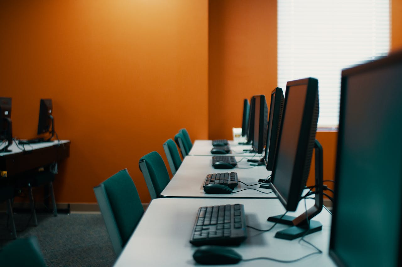 Modern computers with keyboards and mice placed in row on table with chairs  in contemporary workplace with orange walls